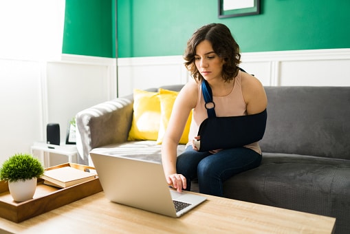 Young woman with arm in sling, seated on couch using a laptop