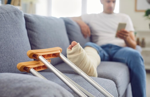 Close up shot of a man broken and bandaged leg resting on a sofa at home, with crutches nearby.
