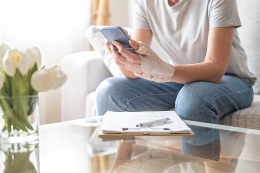 Adult woman sitting on a sofa at home wearing a thumb orthotic splint on her hand while browsing a mobile phone, with paperwork and a pen on the coffee table